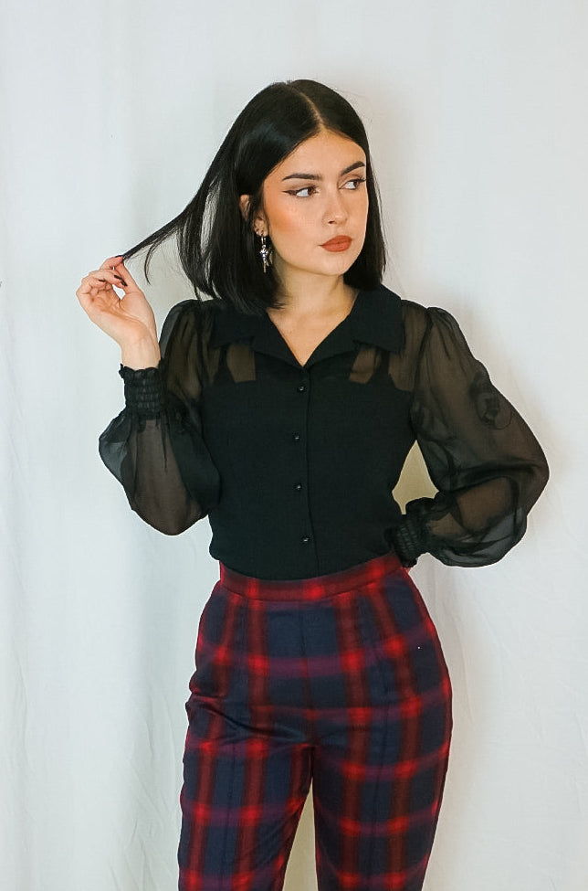 Woman wearing a black sheer blouse and red plaid pants against a white curtain background